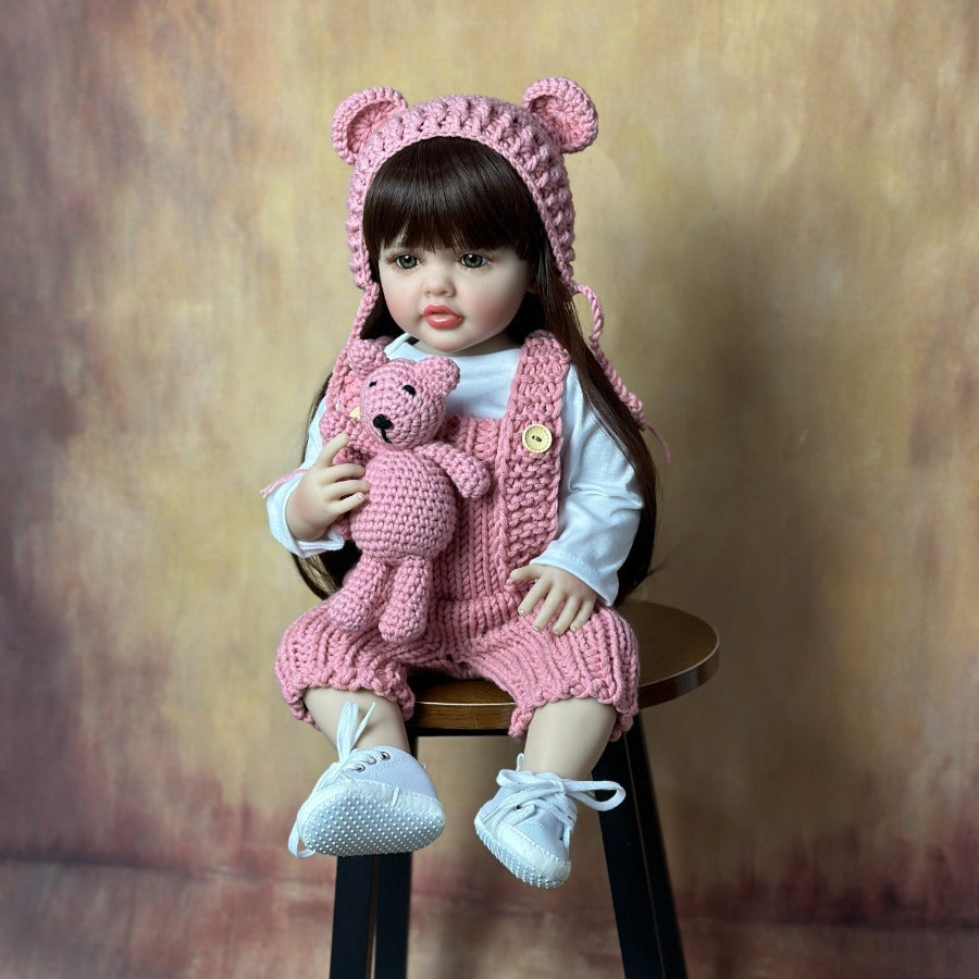 Child wearing a pink bear hat and outfit, holding a pink teddy bear, sitting on a wooden stool against a brown background.
